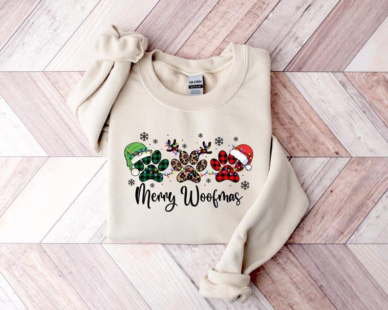May include: A cream-coloured sweatshirt with a Christmas-themed design featuring three paw prints wearing Santa hats. The paw prints are decorated with green and red plaid, leopard print, and green and white plaid. The text "Merry Woofmas" is printed below the paw prints.