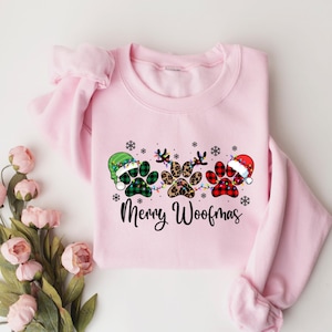 May include: Pink Christmas sweatshirt featuring a design of three paw prints wearing Santa hats and reindeer antlers. The text "Merry Woofmas" is printed below the paw prints.