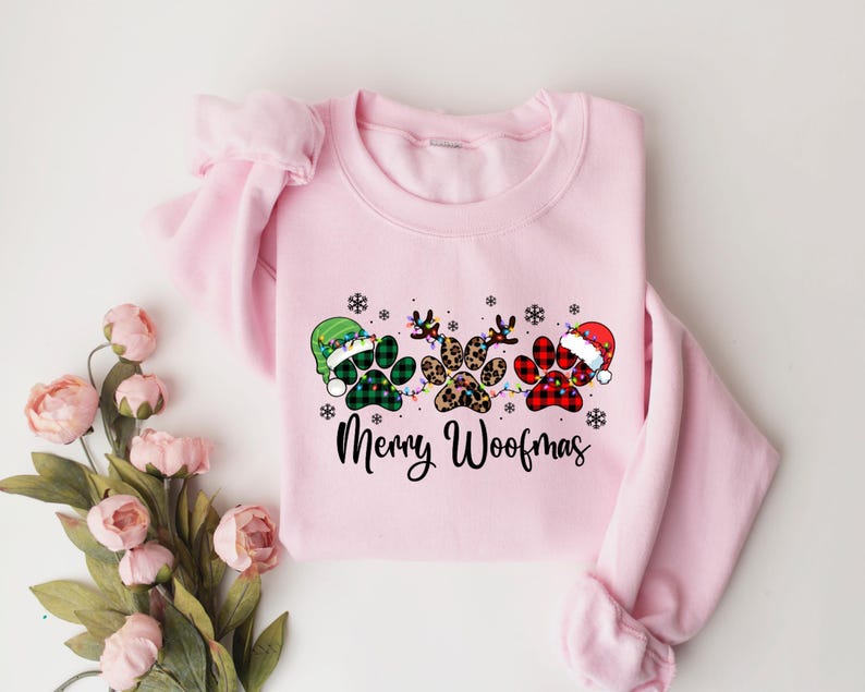 May include: Pink Christmas sweatshirt featuring a design of three paw prints wearing Santa hats and reindeer antlers. The text "Merry Woofmas" is printed below the paw prints.