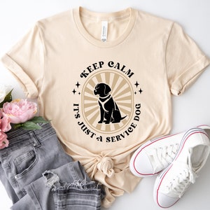 Peut inclure: Un t-shirt beige avec un graphique noir et blanc qui dit "Keep Calm It's Just a Service Dog" avec une silhouette noire d'un chien au centre.