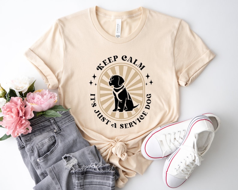 Peut inclure: Un t-shirt beige avec un graphique noir et blanc qui dit "Keep Calm It's Just a Service Dog" avec une silhouette noire d'un chien au centre.