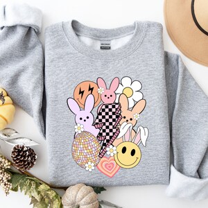 May include: A grey sweatshirt featuring a colourful graphic print of bunnies, a smiley face, a lightning bolt, a daisy and a disco ball. The graphic is in a retro style.