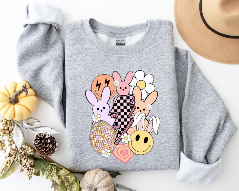 May include: A grey sweatshirt featuring a colourful graphic print of bunnies, a smiley face, a lightning bolt, a daisy and a disco ball. The graphic is in a retro style.
