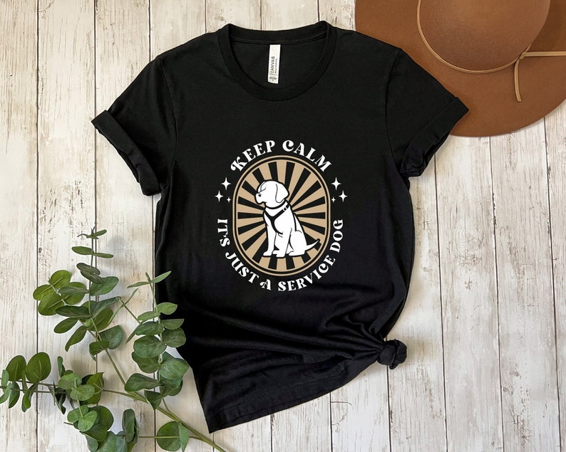 Peut inclure: T-shirt noir avec un motif graphique blanc repr&eacute;sentant un chien portant un gilet de chien d'assistance. Le motif graphique comprend le texte "Keep Calm It's Just a Service Dog" dans un style r&eacute;tro.