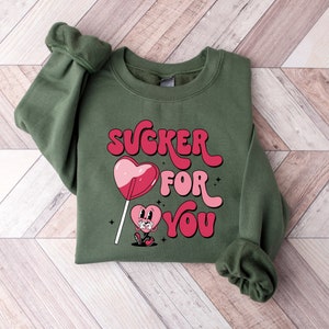 K&ouml;nnte beinhalten: Gr&uuml;ner Pullover mit einem pinkfarbenen und roten Herz-Lollipop-Motiv und dem Text "SUCKER FOR YOU" in einer Retro-Schrift.