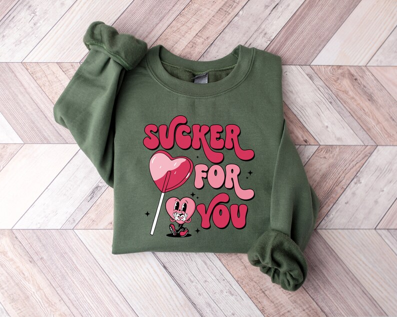 K&ouml;nnte beinhalten: Gr&uuml;ner Pullover mit einem pinkfarbenen und roten Herz-Lollipop-Motiv und dem Text "SUCKER FOR YOU" in einer Retro-Schrift.
