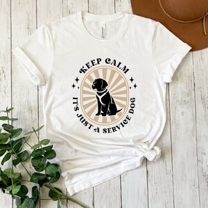 Peut inclure: T-shirt blanc avec un graphique noir et beige qui dit "Keep Calm It's Just a Service Dog" avec une silhouette noire d'un chien portant un gilet de service.