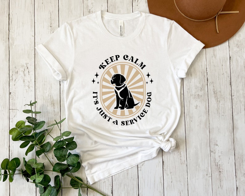 Peut inclure: T-shirt blanc avec un graphique noir et beige qui dit "Keep Calm It's Just a Service Dog" avec une silhouette noire d'un chien portant un gilet de service.