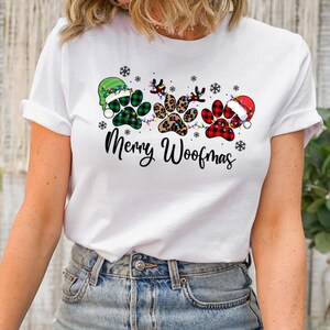 May include: White t-shirt with a Christmas-themed design featuring three paw prints with different patterns, a string of lights, and the text "Merry Woofmas".