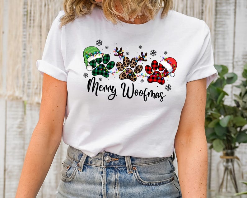 May include: White t-shirt with a Christmas-themed design featuring three paw prints with different patterns, a string of lights, and the text "Merry Woofmas".