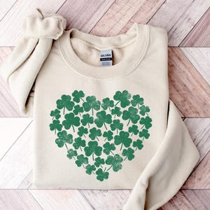 May include: A cream-colored sweatshirt with a green shamrock heart design. The shamrocks are arranged in a heart shape and have a distressed look.