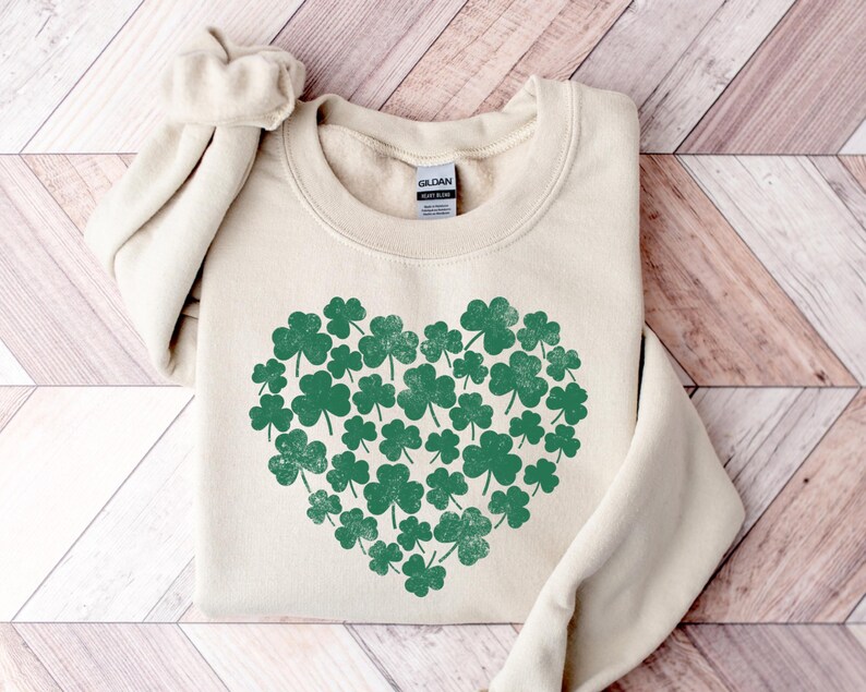May include: A cream-colored sweatshirt with a green shamrock heart design. The shamrocks are arranged in a heart shape and have a distressed look.