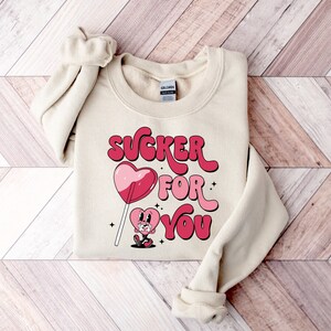 K&ouml;nnte beinhalten: Ein cremefarbener Pullover mit dem Text "SUCKER FOR YOU" in rosa und roten Buchstaben. Ein Cartoon-Herz mit einem Lutscher befindet sich in der Mitte des Textes.