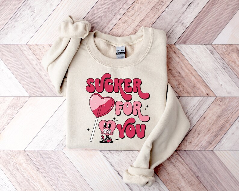 K&ouml;nnte beinhalten: Ein cremefarbener Pullover mit dem Text "SUCKER FOR YOU" in rosa und roten Buchstaben. Ein Cartoon-Herz mit einem Lutscher befindet sich in der Mitte des Textes.