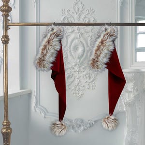 Puede incluir: Dos gorros de Papá Noel de color rojo intenso con ribete de piel sintética. Cada gorro tiene un pompón esponjoso. Cuelgan de una barra dorada para ropa contra una pared blanca.