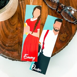 May include: Two illustrated bookmarks with blue backgrounds. One bookmark features a woman with long red hair and a red dress holding a book. The bookmark is labeled "Emmy". The other bookmark features a man with brown hair and a white shirt with red suspenders. The bookmark is labeled "Owen".