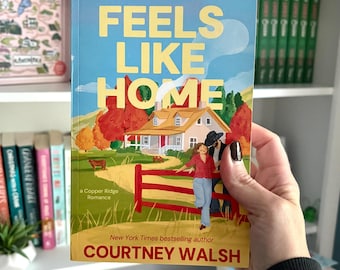 May include: A paperback book titled Feels Like Home by Courtney Walsh. The cover features a light blue sky, a house with a red roof, autumn trees, a red fence, and two figures standing together. The text reads Feels Like Home and Courtney Walsh.