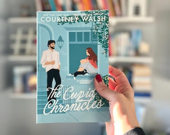 May include: A blue book cover with the title "The Cupid Chronicles" in white script. The book cover features a whimsical illustration of a man and woman standing on a porch with a cat.