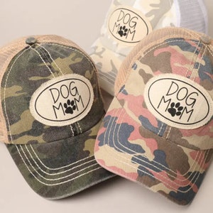 May include: Three camouflage trucker hats with a brown oval patch that says "Dog Mom" in black text. The hats have a mesh back and a brown brim.