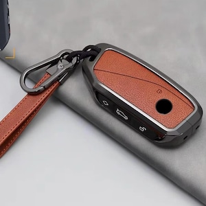 Può includere: Una custodia per chiave auto con inserto in pelle marrone testurizzata e cornice in metallo scuro. Include un cinturino in pelle marrone abbinato fissato a un moschettone in metallo scuro. Il lato mostra icone bianche per le funzioni del veicolo.