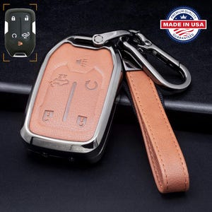 May include: A car key fob cover with a brown leather insert and a silver-colored metal frame. The fob has a matching brown leather strap and a silver-colored carabiner clip. The key fob cover is marked "MADE IN USA".