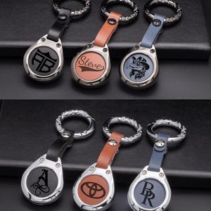 Può includere: Una collezione di portachiavi personalizzati con vari design. Ogni portachiavi presenta un ciondolo metallico a forma di goccia con un cinturino in pelle e un anello decorativo. I ciondoli mostrano iniziali, nomi e grafiche in nero e marrone.