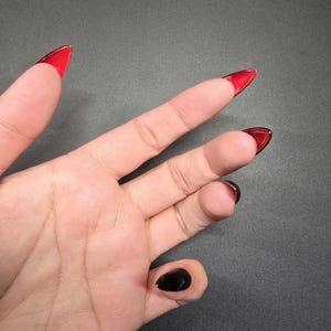 Puede incluir: Una mano con uñas postizas largas, puntiagudas, rojas y negras. Las uñas son de color rojo oscuro con una punta negra.