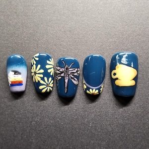 Puede incluir: Cinco diseños de esmalte de uñas azul con diferentes patrones. Los diseños incluyen un barco, margaritas, una libélula, un borde de margaritas y una taza de té.