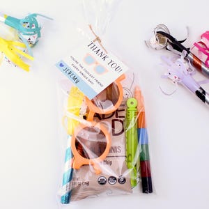 May include: A clear plastic bag filled with party favors for a child's birthday party. The bag contains a pair of orange plastic sunglasses, a small bag of chocolate chips, a pack of crayons, and a thank you tag that says "Thank You! You're the coolest friend every single day! Jeremy".