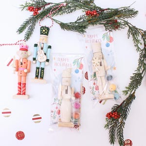 May include: Two painted wooden nutcracker ornaments and two DIY nutcracker kits in clear plastic packaging. The kits include a plain wooden nutcracker, a small paintbrush, and three small paint pots, all labeled with the text Happy Holidays.