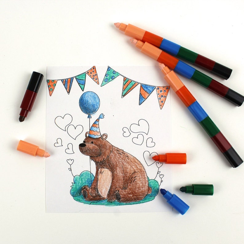 May include: A coloring page featuring a brown bear wearing a party hat and holding a blue balloon. The bear is sitting on a green patch of grass with a string of colorful flags above. The page is surrounded by colorful markers.