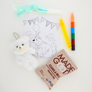 May include: A coloring page featuring a bear wearing a party hat and a balloon, surrounded by hearts. A white teddy bear, a yellow and blue bubble wand, a multi-colored marker, and a bag of chocolate chip granola minis are also included.