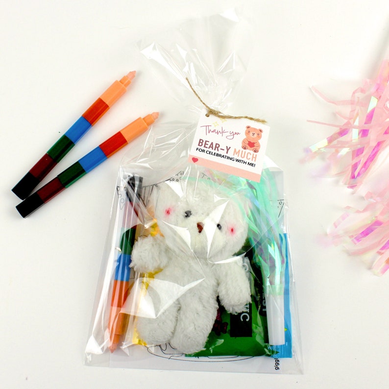 May include: A clear plastic bag filled with party favors for a teddy bear themed party. The bag contains a small white teddy bear, crayons, a coloring page, and a thank you tag that reads "Thank you BEAR-Y MUCH for celebrating with me!"