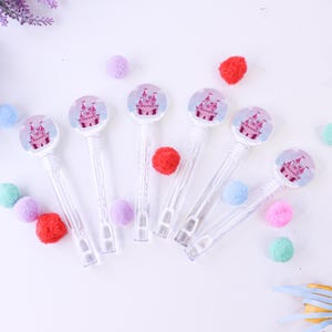 May include: Several clear bubble wands with a pink castle design on the top, surrounded by colorful pom-poms. The wands are arranged on a white surface, creating a playful and whimsical scene.