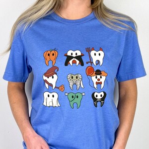 Pode incluir: Uma camiseta azul claro com um gráfico de nove dentes vestidos com diferentes fantasias de Halloween. Os dentes estão vestidos como uma abóbora, um vampiro, um diabo, uma bruxa, uma múmia, um palhaço, um fantasma, um zumbi e um esqueleto.