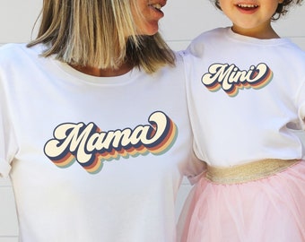 May include: A white t-shirt with the word "Mama" in a retro rainbow font. A child is wearing a white t-shirt with the word "Mini" in the same font. The child is wearing a pink tulle skirt.