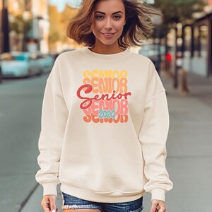 Könnte beinhalten: Ein weißer Pullover mit einer Retro-Grafik, auf der "Senior 2024" in bunten Buchstaben steht.