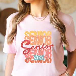 Könnte beinhalten: Rosa T-Shirt mit einer Retro-Grafik, auf der "Senior 2024" in einem Regenbogen aus Farben steht.