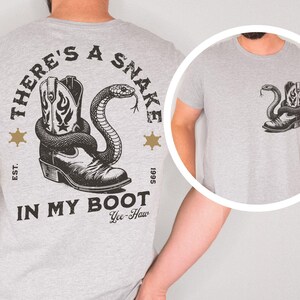 Puede incluir: Camiseta gris claro con un gr&aacute;fico de una bota de vaquero y una serpiente. El texto "THERE'S A SNAKE IN MY BOOT" est&aacute; impreso arriba y abajo del gr&aacute;fico, con "Yee-Haw" debajo. Dos estrellas doradas a cada lado.