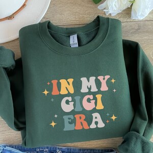 Puede incluir: Una sudadera verde oscuro con el texto "IN MY GIGI ERA" en letras retro de colores. El texto est&aacute; rodeado de peque&ntilde;as estrellas doradas.
