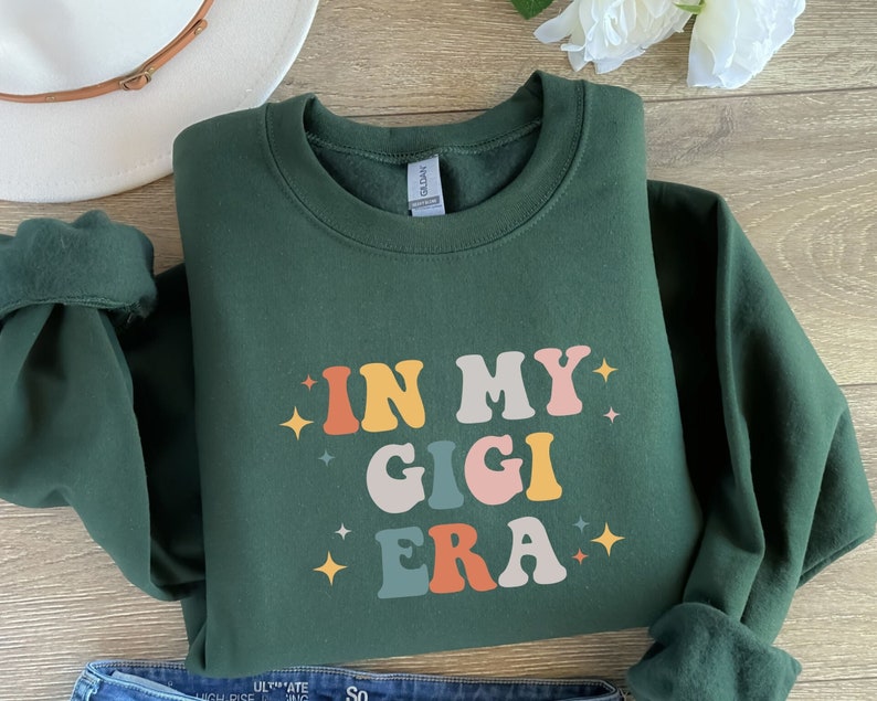 Puede incluir: Una sudadera verde oscuro con el texto "IN MY GIGI ERA" en letras retro de colores. El texto est&aacute; rodeado de peque&ntilde;as estrellas doradas.