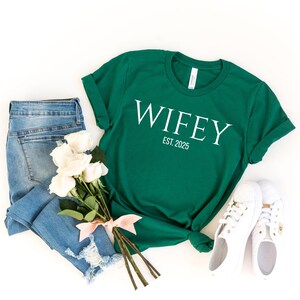 May include: A green t-shirt with the word "WIFEY" in white, and the text "EST. 2025". The shirt is paired with light blue jeans, white sneakers, and a bouquet of white roses tied with a pink ribbon.