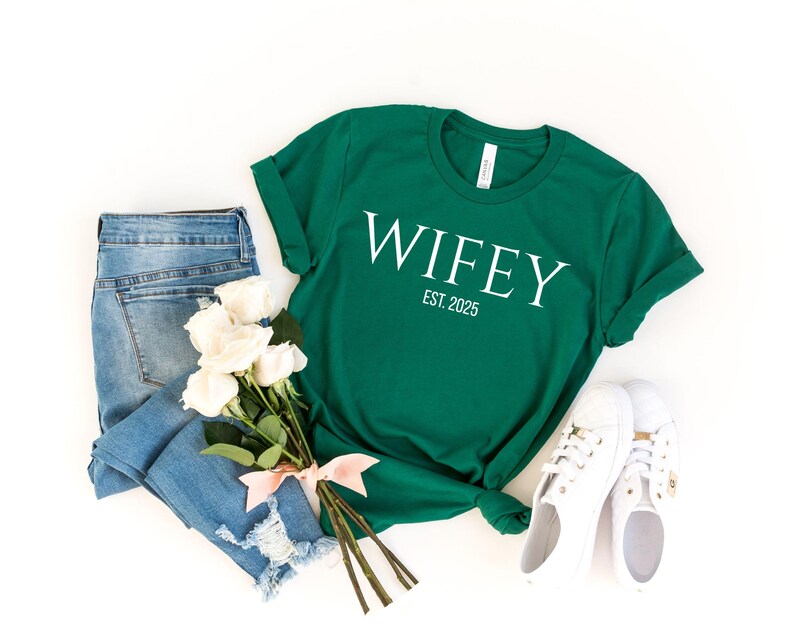 May include: A green t-shirt with the word "WIFEY" in white, and the text "EST. 2025". The shirt is paired with light blue jeans, white sneakers, and a bouquet of white roses tied with a pink ribbon.
