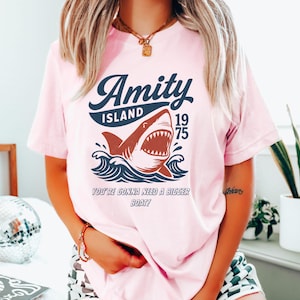 以下が含まれることがあります： ピンク色のTシャツ。波から現れるサメのグラフィック、"Amity Island 1975"の文字、そして"You're Gonna Need a Bigger Boat!"というフレーズがネイビーと赤で描かれています。