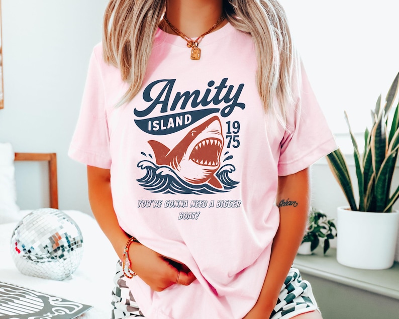 以下が含まれることがあります： ピンク色のTシャツ。波から現れるサメのグラフィック、"Amity Island 1975"の文字、そして"You're Gonna Need a Bigger Boat!"というフレーズがネイビーと赤で描かれています。