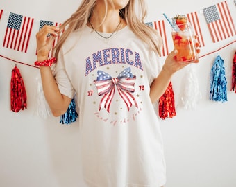 May include: Cream-colored t-shirt with a patriotic design. The shirt features the word "AMERICA" in blue and red stars, a bow with the American flag design, and the numbers "17" and "76". The phrase "land of the free" is written below the bow.