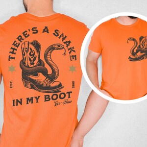 Puede incluir: Camiseta naranja con un gr&aacute;fico negro de una bota de vaquero y una serpiente. El texto "THERE'S A SNAKE IN MY BOOT" est&aacute; impreso en la espalda. Una versi&oacute;n m&aacute;s peque&ntilde;a del gr&aacute;fico est&aacute; en el &aacute;rea del bolsillo delantero. La camiseta tambi&eacute;n tiene el texto "Yee-Haw".