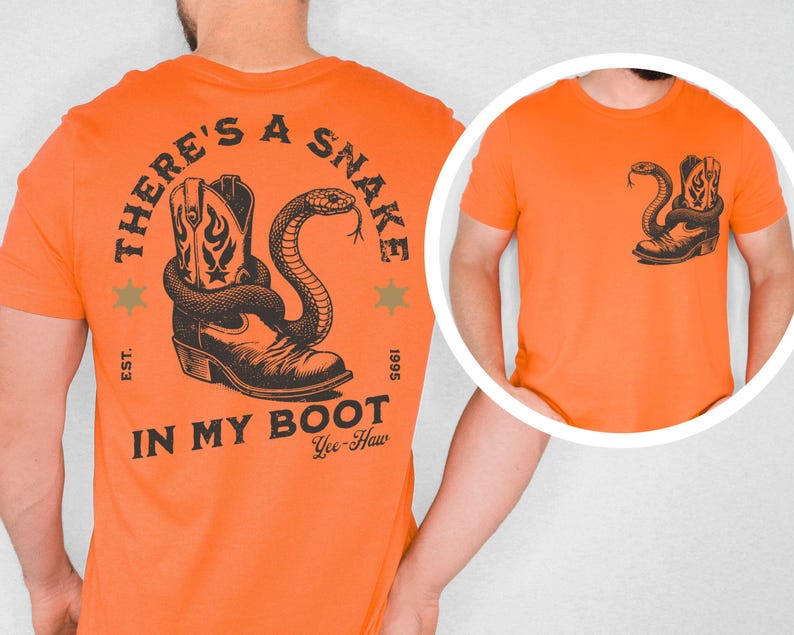 Puede incluir: Camiseta naranja con un gr&aacute;fico negro de una bota de vaquero y una serpiente. El texto "THERE'S A SNAKE IN MY BOOT" est&aacute; impreso en la espalda. Una versi&oacute;n m&aacute;s peque&ntilde;a del gr&aacute;fico est&aacute; en el &aacute;rea del bolsillo delantero. La camiseta tambi&eacute;n tiene el texto "Yee-Haw".