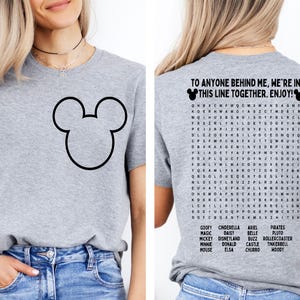 K&ouml;nnte beinhalten: Ein hellgraues T-Shirt mit einer schwarzen Umrisszeichnung von Mickey Mouse's Kopf auf der Vorderseite. Auf der R&uuml;ckseite steht der Text "TO ANYONE BEHIND ME, WE'RE IN THIS LINE TOGETHER. ENJOY!" und ein Disney-Wortsuchr&auml;tsel.