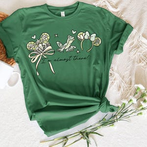 Puede incluir: Camiseta verde con un dise&ntilde;o caprichoso que presenta una rana, un lazo y elementos florales. El texto "I'm almost there!" est&aacute; impreso debajo del gr&aacute;fico. La camiseta est&aacute; anudada en la parte inferior.